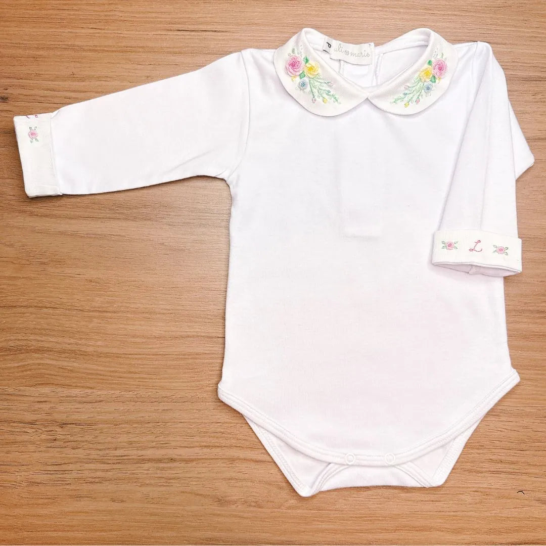 body martina branco em malha de algodão com gola redonda bordada com flores coloridas, sobre fundo de madeira