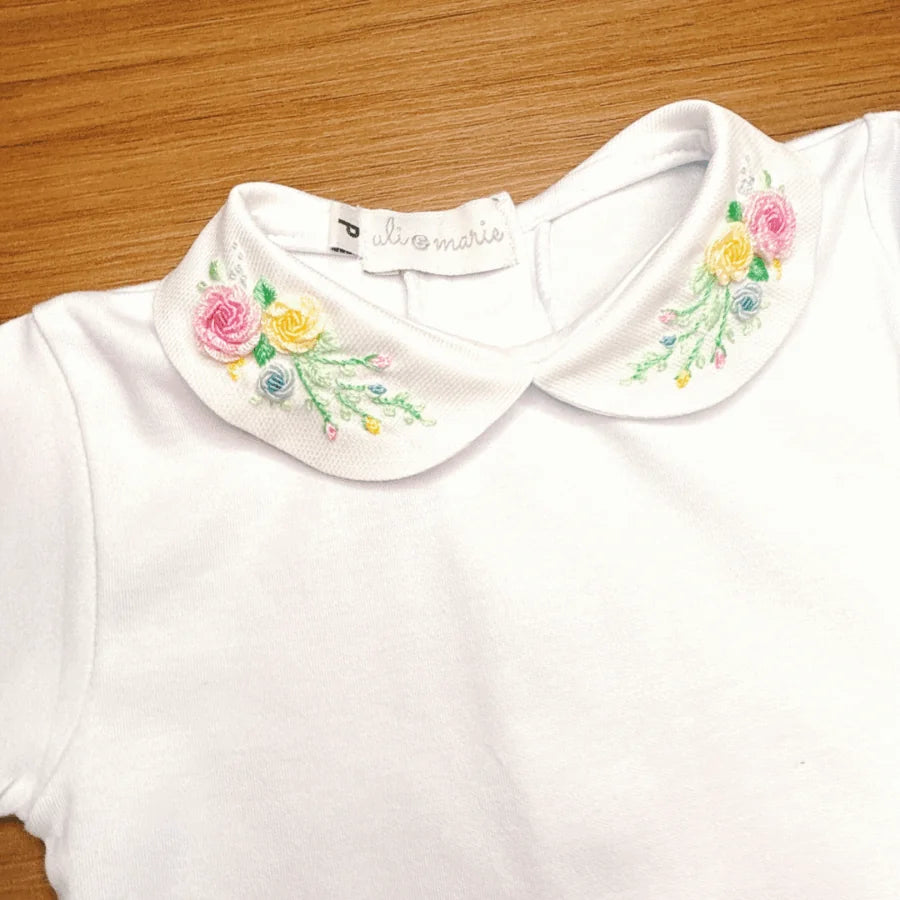 body martina branco em malha de algodão com gola redonda bordada com flores coloridas, sobre fundo de madeira