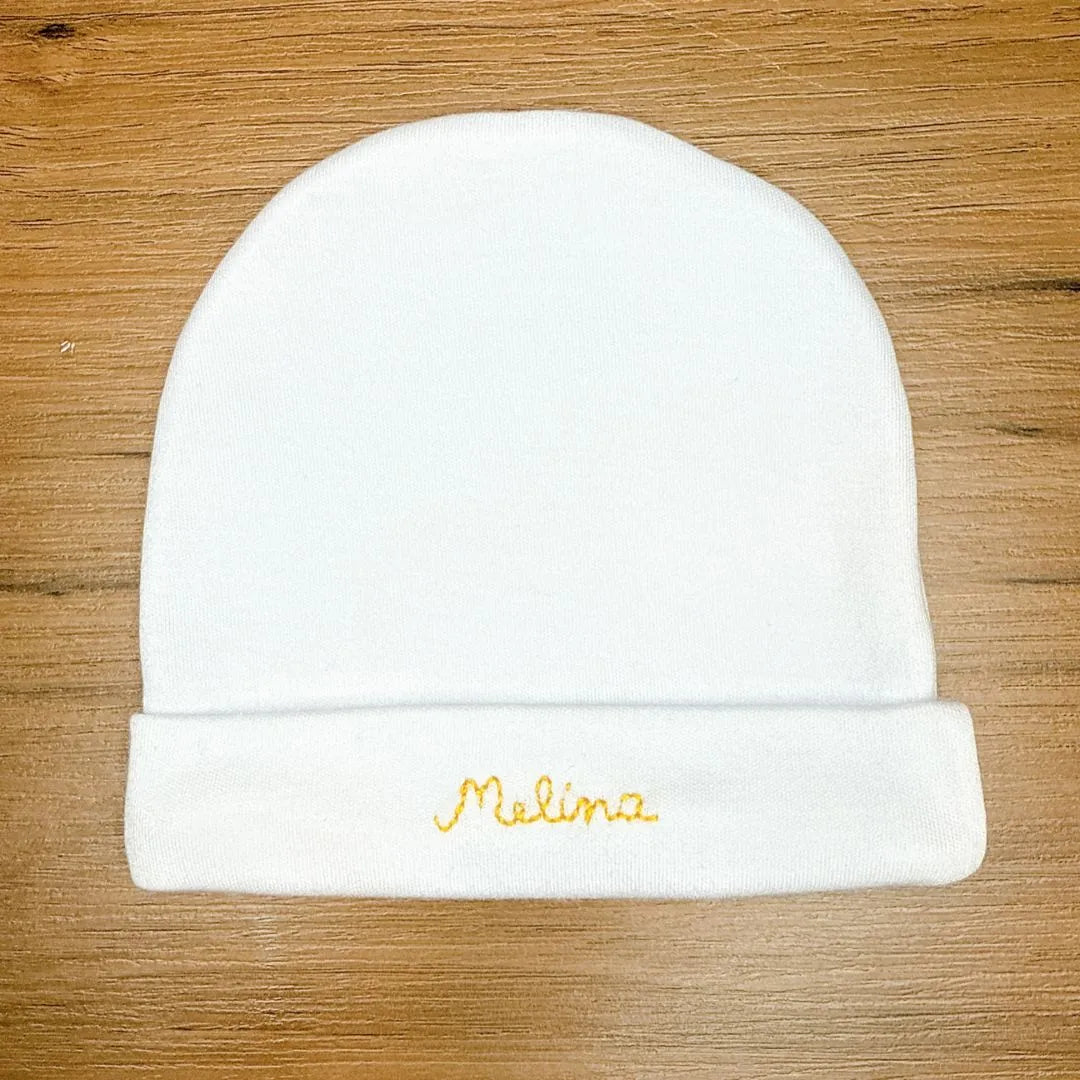 touca de malha branca do kit maternidade, dobrada na parte inferior para mostrar o nome 'melina' bordado em amarelo