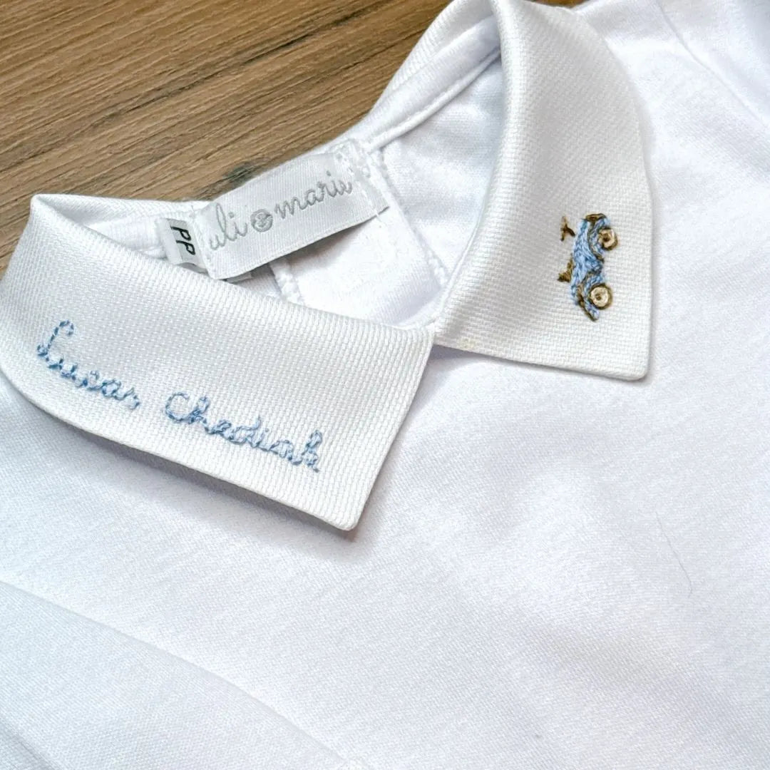 close-up da gola do body com bordado de carrinho azul e acabamento delicado, tecido branco macio em destaque